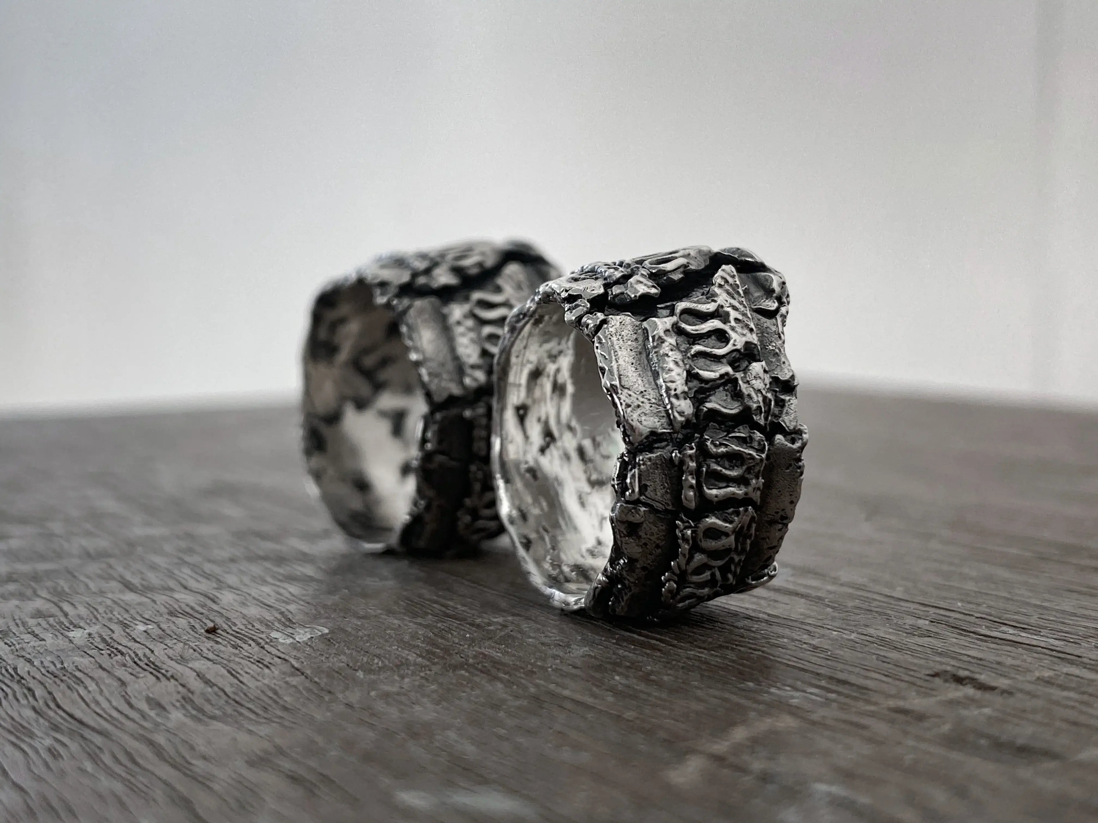 Two textured silver rings with a hammered, organic surface finish, displayed on a dark wooden surface against a neutral light gray background.