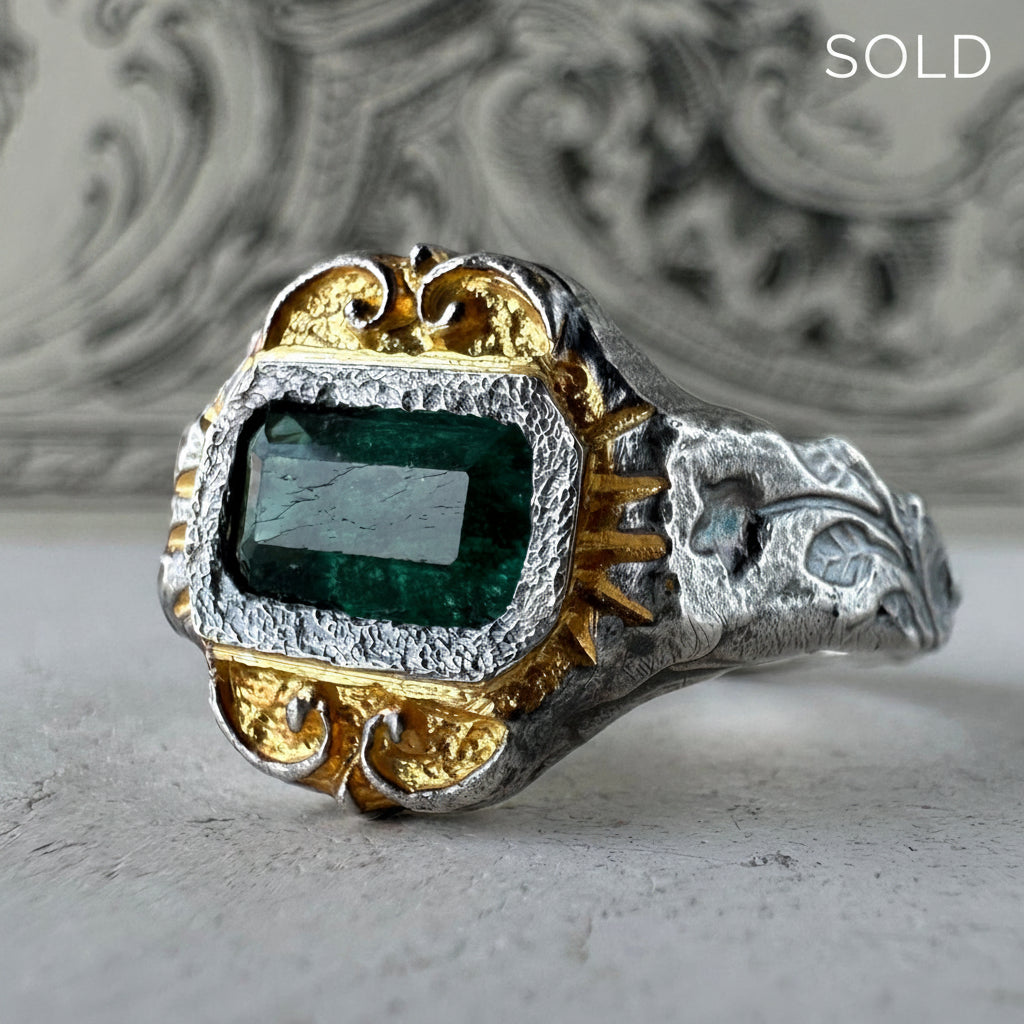 A two-tone silver and gold ring featuring a rectangular emerald-cut green gemstone, set against a blurred gray background with a 'SOLD' text overlay in the top right corner.