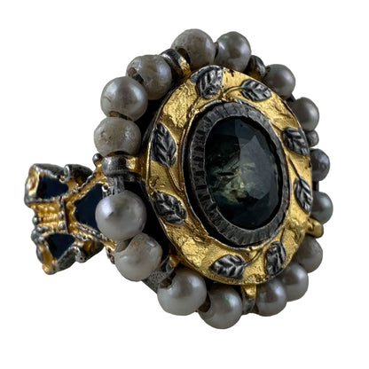 An antique-style ring featuring a round dark green faceted gemstone in a gold and silver-toned setting, surrounded by a border of gray pearls.